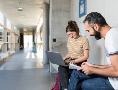Zwei Studierende sitzen in einem hellen Gang des Geisteswissenschaftlichen Zentrums der Universität Leipzig. Sie beschäftigen sich mit einem Laptop und machen sich Notizen. Sie machen einen fröhlichen Eindruck.