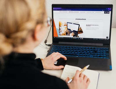 Farbfoto: Eine junge Frau sitzt mit dem Rücken zur Kamer vor einem Laptop und recherchiert auf der Website der Universität Leipzig nach passenden Studiengängen.